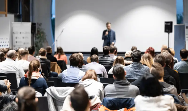 Audience seated in a conference hall listening to a speaker standing in front of a large projection screen.