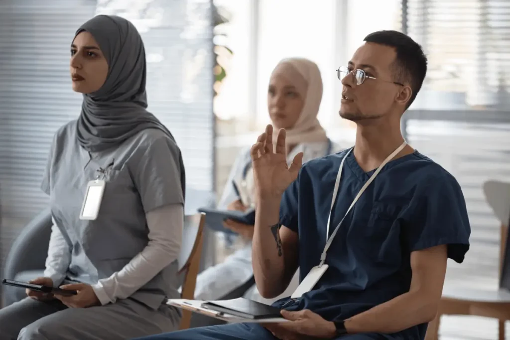 Medical professionals in training sit in a classroom, with one participant raising his hand to ask a question.