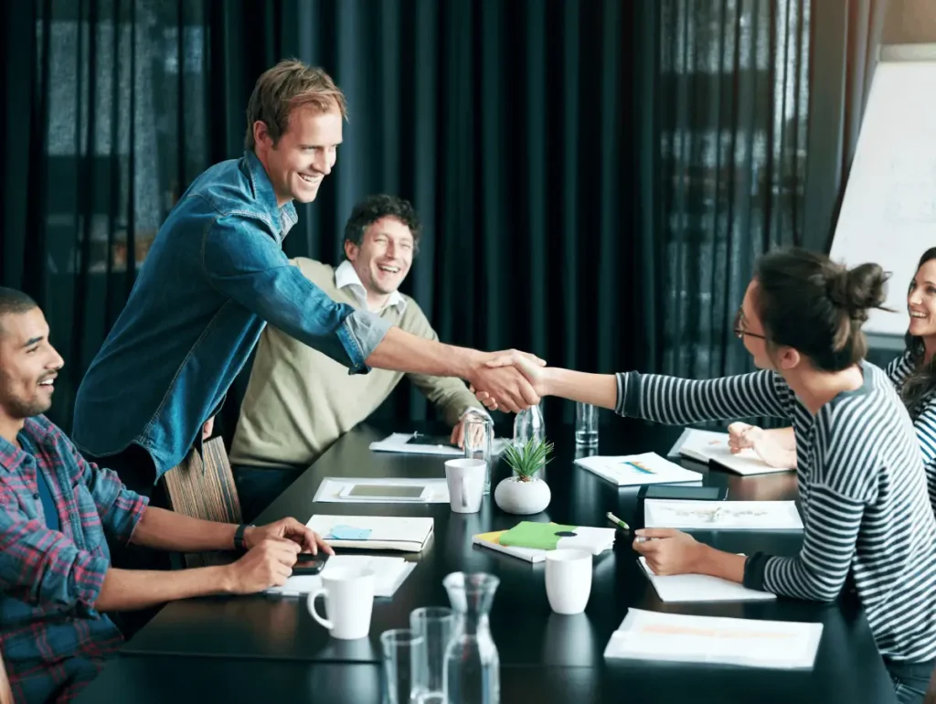 A group of colleagues sitting around a table in a meeting room, smiling as two of them shake hands across the table.