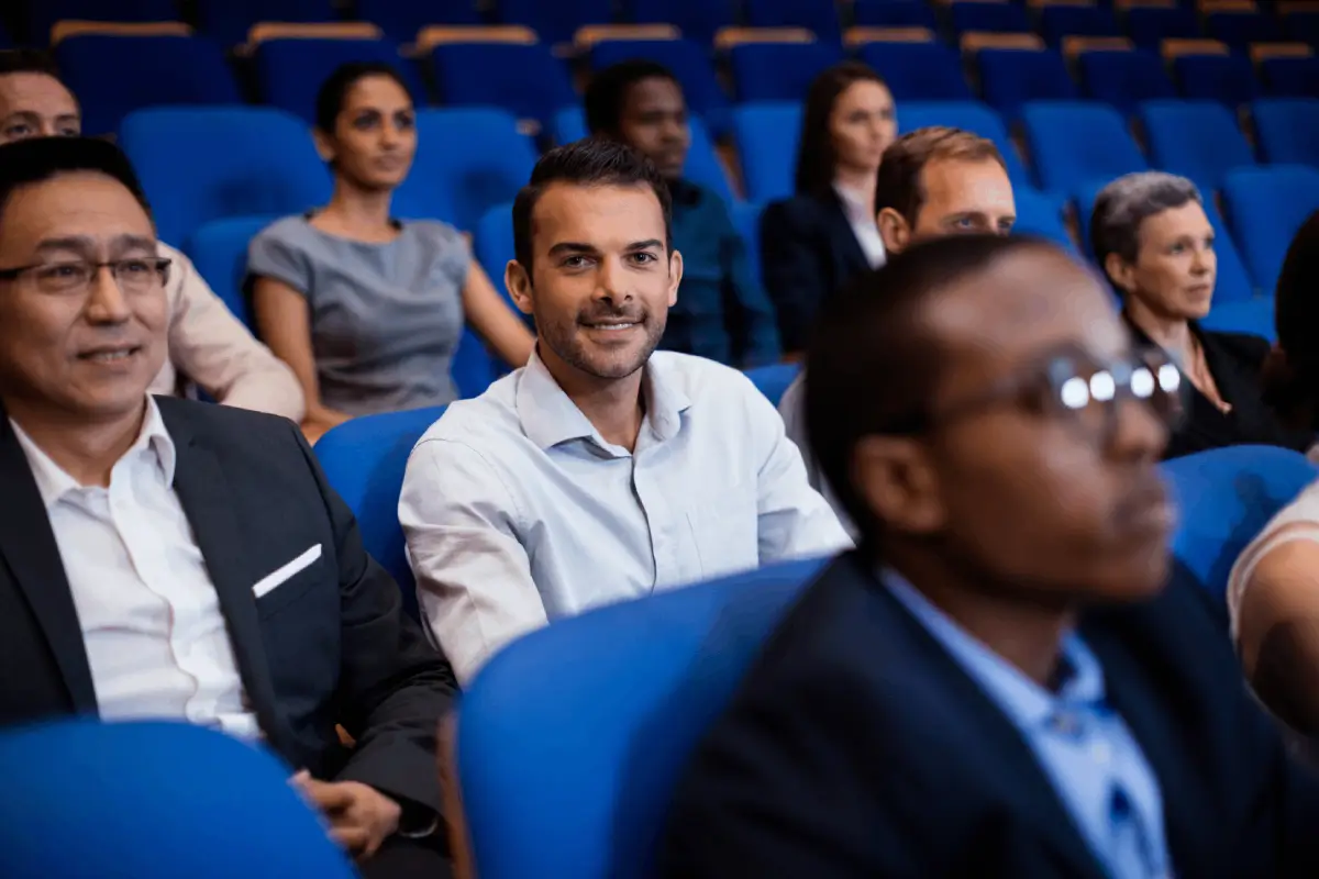 People of different ages and ethnic backgrounds are seated in blue chairs at a conference or seminar. A man in the middle row is smiling directly at the camera.
