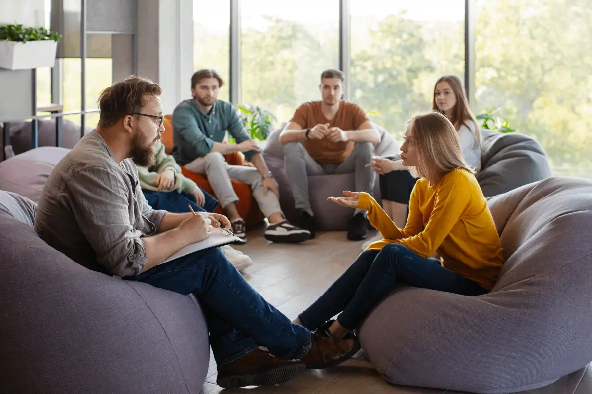 A small group therapy session where people sit on bean bag chairs in a bright room. A woman in a yellow sweater is speaking while a man with a notepad listens attentively.