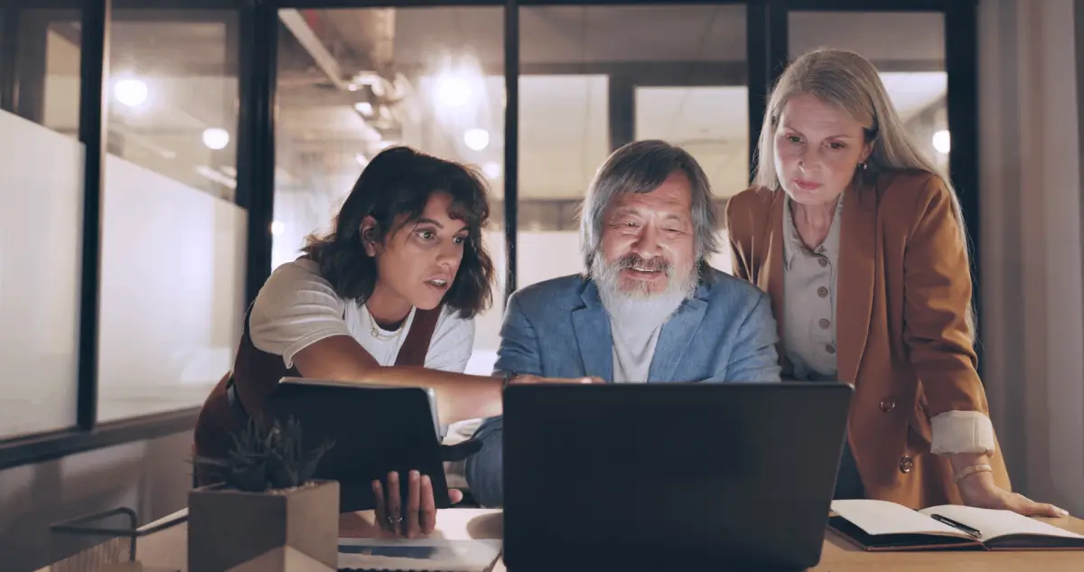 Three colleagues are gathered around a laptop, discussing something on the screen with focus and collaboration.