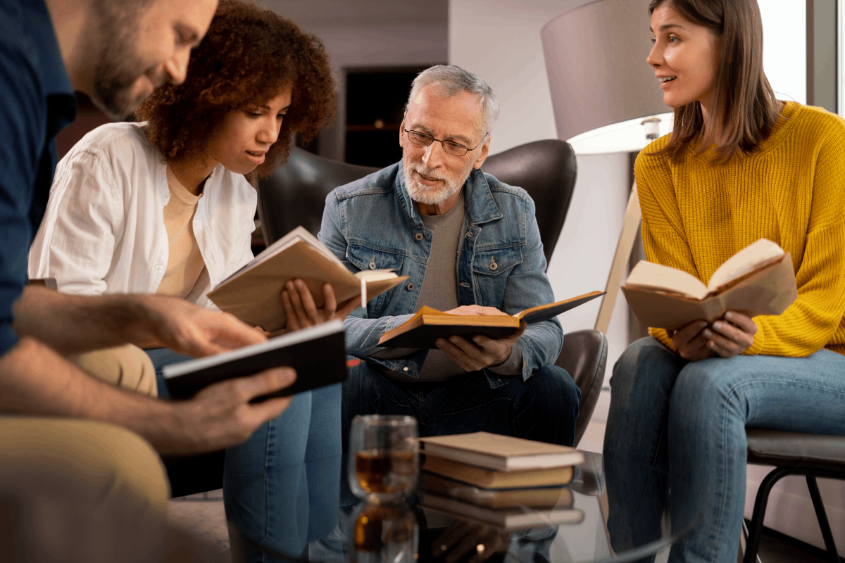 A diverse group of people sitting together in a cozy room, holding and reading books, while engaging in discussion. An older man in a denim jacket is looking at a young woman explaining from her book.