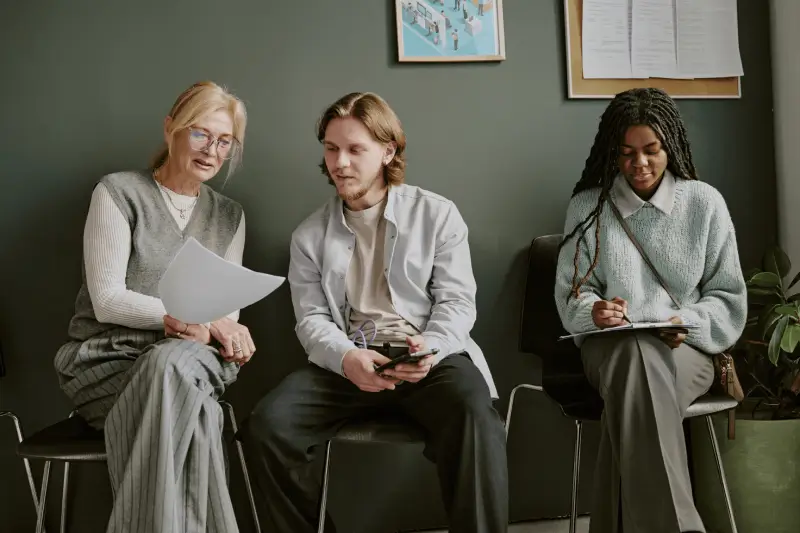 Three people are sitting in chairs against a wall, reviewing documents and taking notes together in a casual setting.
