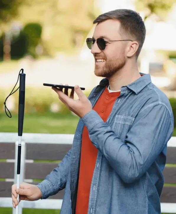 A blind man wearing sunglasses smiles while holding a smartphone with voice command, standing outdoors with a white cane in his other hand.