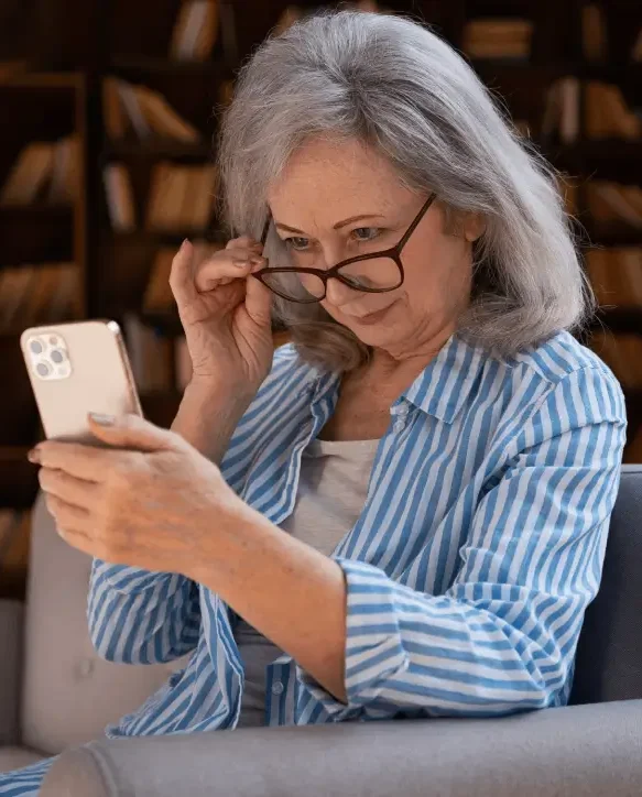 An older woman with gray hair adjusts her glasses while looking closely at a smartphone.