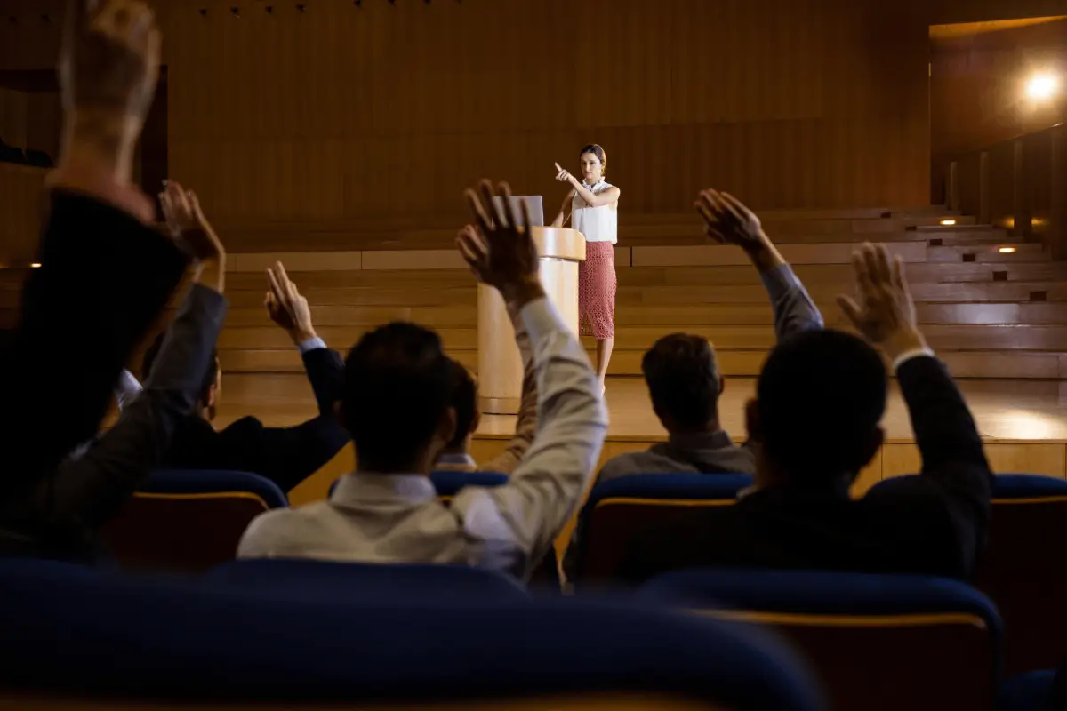 A woman stands at a podium in an auditorium, addressing an audience with several people raising their hands.